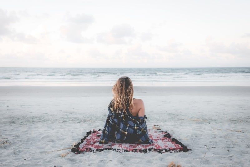 Frau sitzt auf einer Decke am Strand und blickt aufs Meer