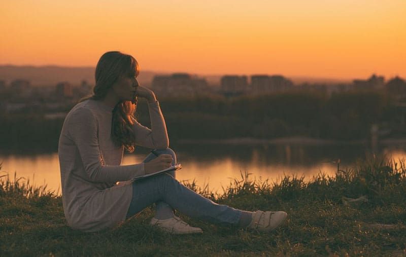 Frau schreibt auf Notizbuch und sitzt bei Sonnenuntergang auf dem Boden in der Nähe eines Gewässers