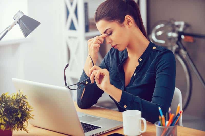 Eine frustrierte junge Frau hält die Augen geschlossen, während sie an ihrem Arbeitsplatz im Büro sitzt