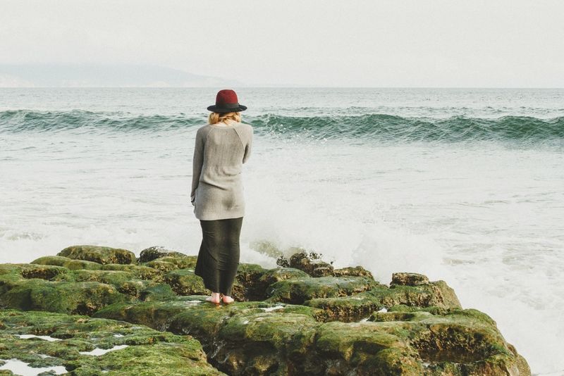 Frau mit Hut steht auf einem Felsen und blickt auf das Meer
