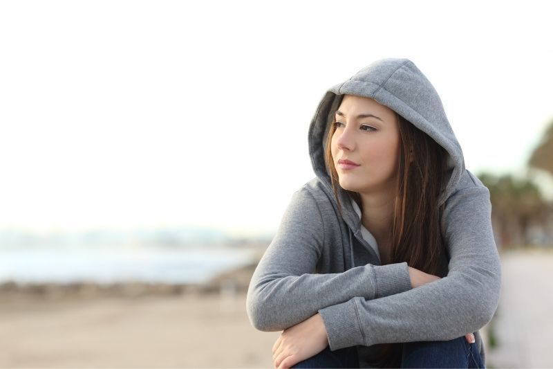 Nachdenkliche Frau auf Kapuzenpullover, die tagsüber am Strand sitzt