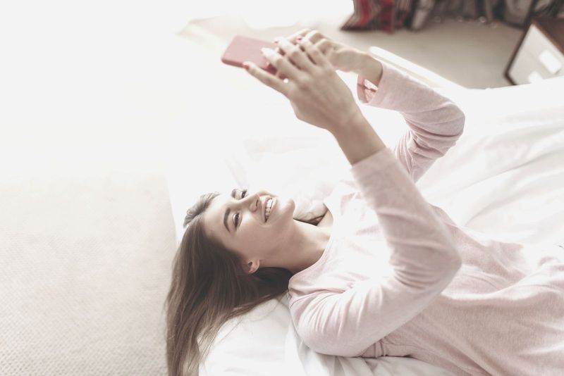 Junge Frau liegt morgens lächelnd im Bett und schaut auf ihr Smartphone