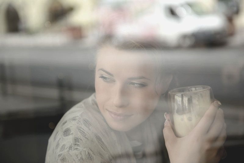 Junge Frau hält ein Glas Frappe hinter dem Fenster und denkt nach