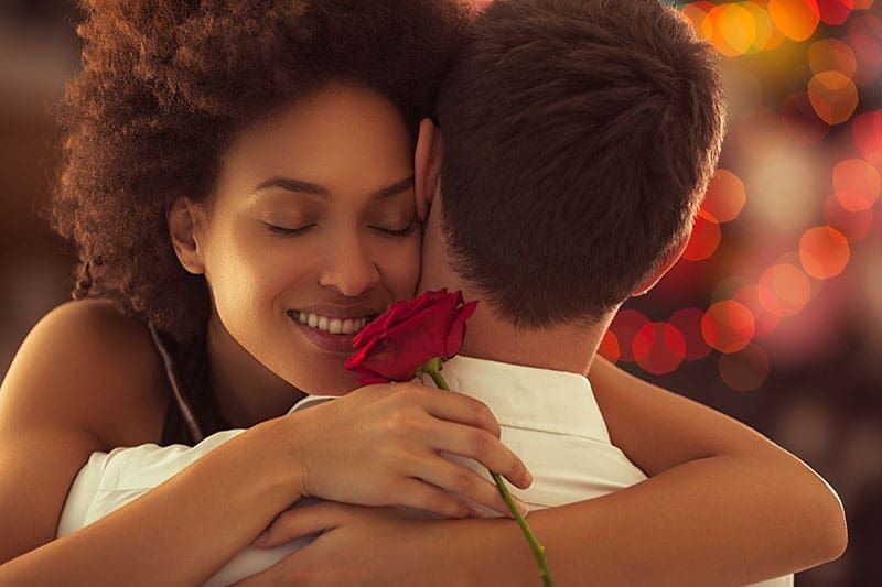 Lächelnde Afrikanerin umarmt ihren Freund und hält die Rose, die sie zum Valentinstag bekommen hat
