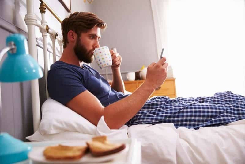 Ein Mann sitzt im Bett, trinkt Kaffee und drückt auf ein Telefon