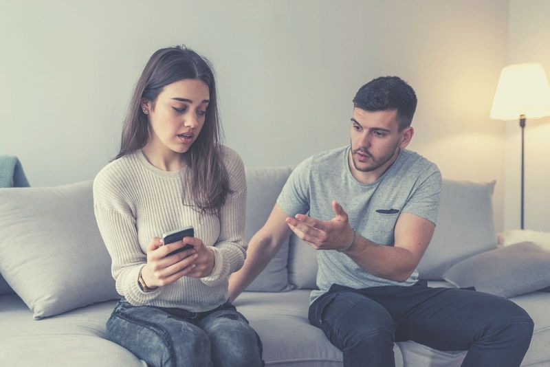 Eifersüchtiger Freund bittet seine Freundin um das Telefon, während er SMS schreibt und neben ihm auf dem Sofa sitzt
