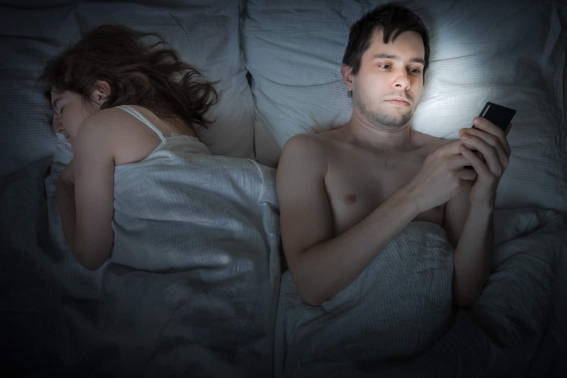 Mann benutzt Smartphone, während er neben Frau im Bett liegt