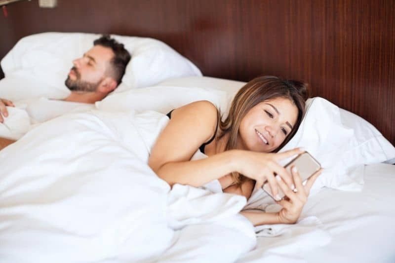 Frau, die neben ihrem schlafenden Ehemann auf dem Bett SMS schreibt