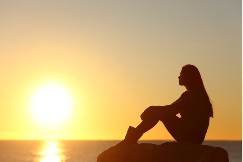 Profil einer Frauensilhouette, die während des Sonnenuntergangs auf einem Felsen in der Nähe des Wassers sitzt