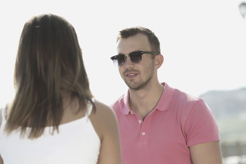 Junger Mann trägt rosa Poloshirt und Sonnenbrille, während er einer Frau im Freien gegenübersteht und mit ihr spricht