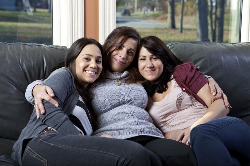 Frau und zwei Töchter sitzen auf der Couch im Wohnzimmer und schauen lächelnd in die Kamera