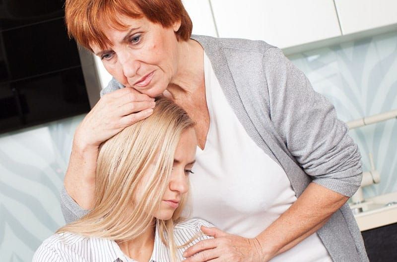 Ältere Frau umarmt eine traurige Frau mit blonden Haaren, die sitzt und sich an ihre Brust lehnt