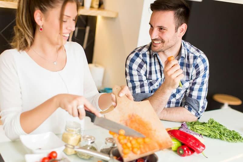 Mann und Frau kochen, während eine Frau ein Messer hält und Gemüse einschenkt