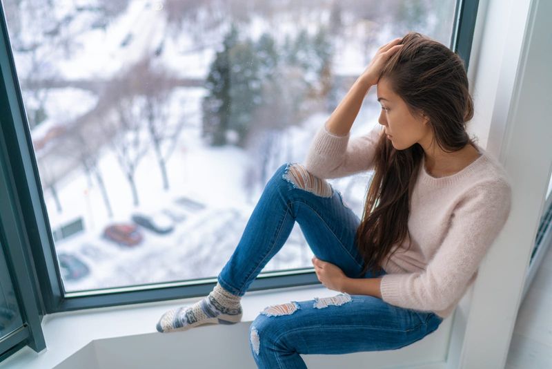 trauriges Mädchen, das einsam am Fenster zu Hause steht und das kalte Wetter betrachtet