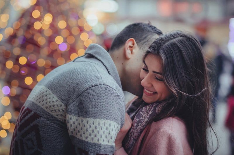 Mann küsst Frau draußen in der Nähe eines großen Weihnachtsbaums auf den Hals