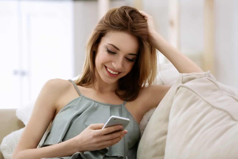 Eine lächelnde Frau sitzt auf der Couch und schaltet das Telefon ein
