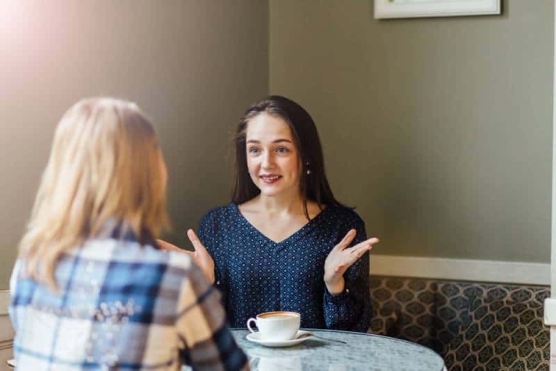 Fröhliche Frau teilt die Neuigkeiten mit ihrer Freundin, während sie in einem Café Cappuccino trinkt