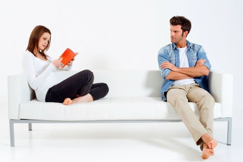 verärgerter Mann, der auf dem Sofa sitzt, während eine Frau am gegenüberliegenden Ende des Sofas ein Buch liest