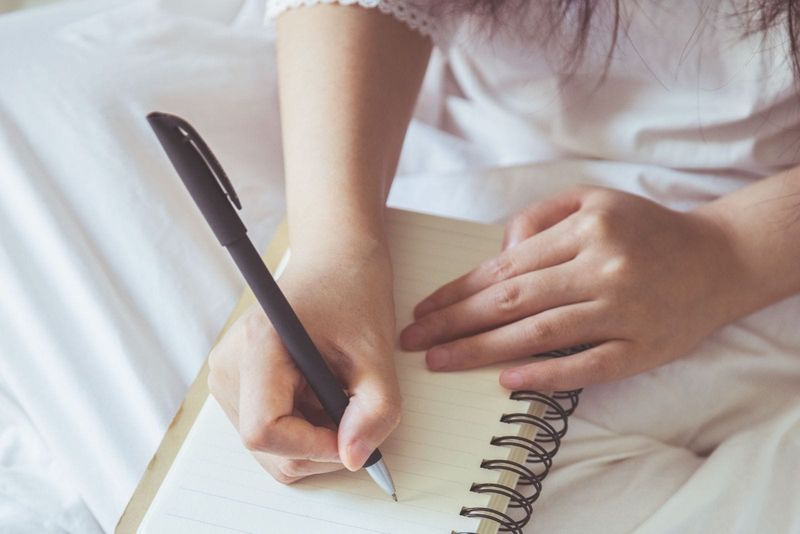 Ausgeschnittenes Foto einer Frau, die im Bett etwas auf ihr Notizbuch schreibt