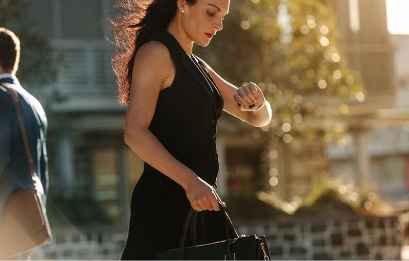 Geschäftsfrau schaut auf ihre Armbanduhr und trägt eine Tasche in einem schwarzen Kleid