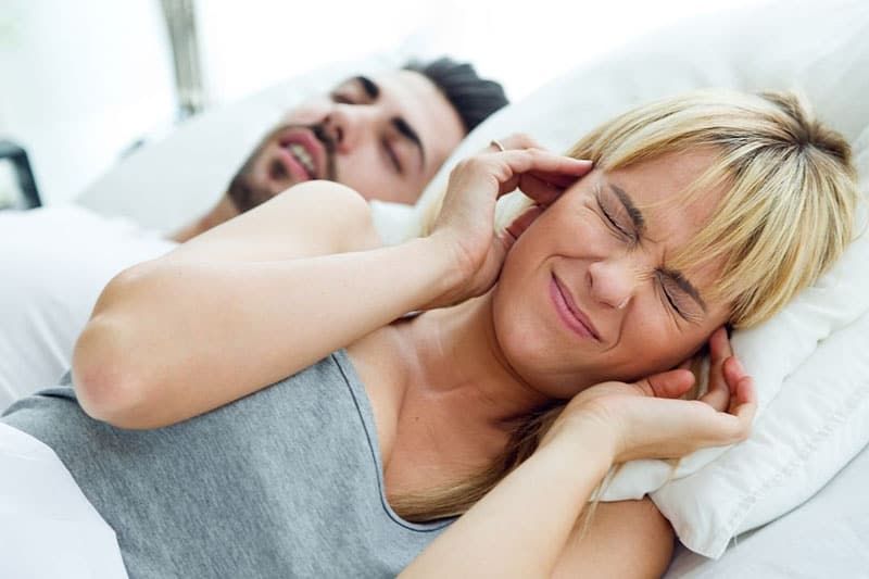 Frau hält sich die Ohren zu und ärgert sich darüber, dass der Mann schnarcht, während er neben ihr schläft
