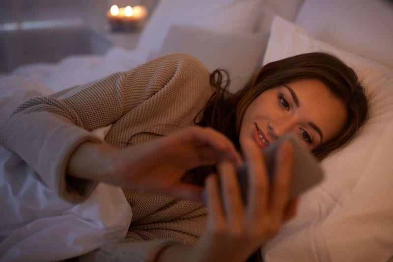 Lächelnde junge Frau, die im Bett auf dem Smartphone SMS schreibt