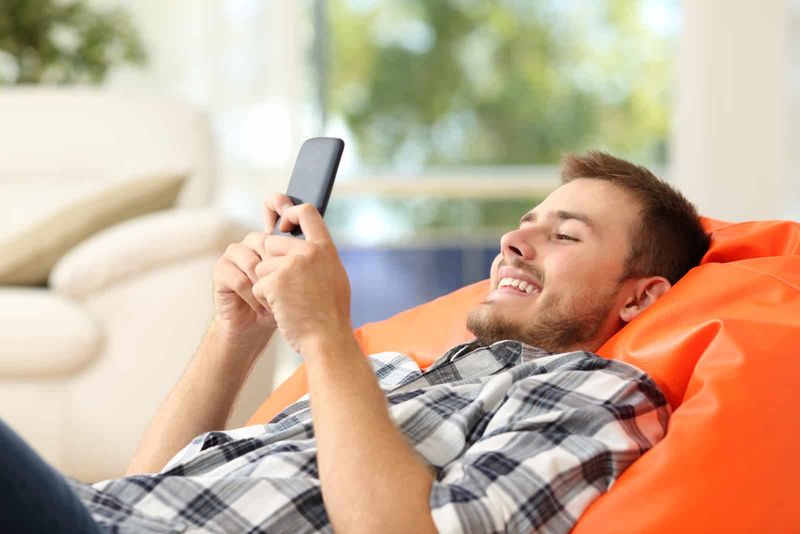 Entspannter Mann mit einem Smartphone, der auf einem orangefarbenen Hocker liegt