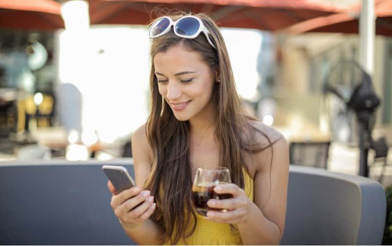 Glückliche Frau, die auf ihrem Telefon mit Weinglas in der anderen Hand SMS schreibt