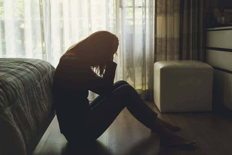 Einsame junge Frau, depressiv und gestresst, sitzt im dunklen Schlafzimmer mit dem Kopf in den Händen