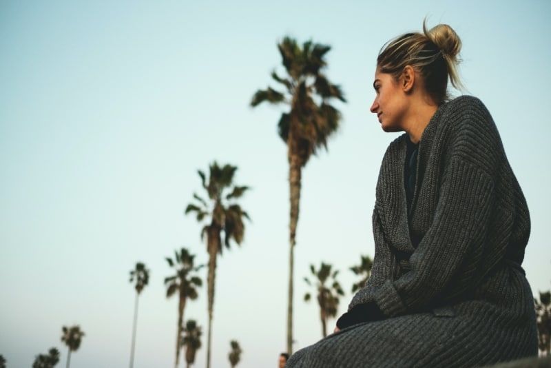 Frau im grauen Pullover sitzt in der Nähe von Kokospalmen im Freien