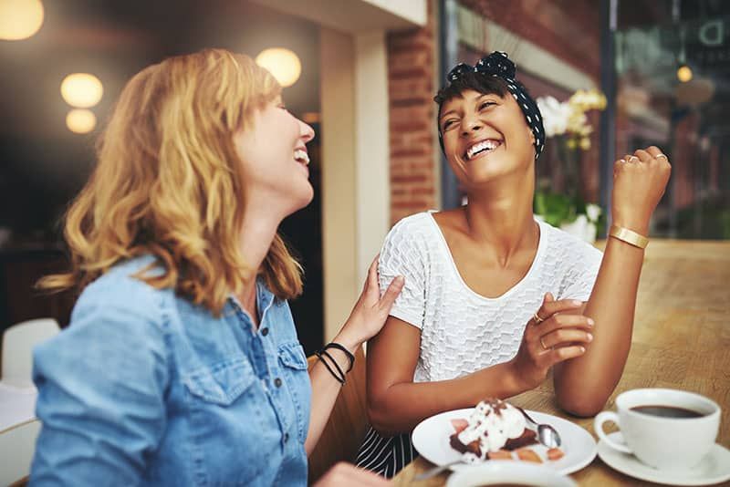 Zwei multiethnische junge Freundinnen genießen gemeinsam Kaffee in einem Restaurant, lachen und scherzen, während sie sich berühren, um ihre Zuneigung zu zeigen