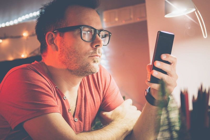 Mann benutzt Smartphone in rotem Hemd und Brille während der Nacht in seinem Heimbüro