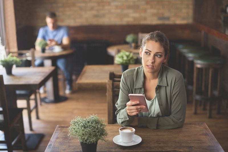 Nachdenkliche Frau, die Textnachrichten im Café sendet, mit einem verschwommenen Mann, der hinten Kaffee trinkt