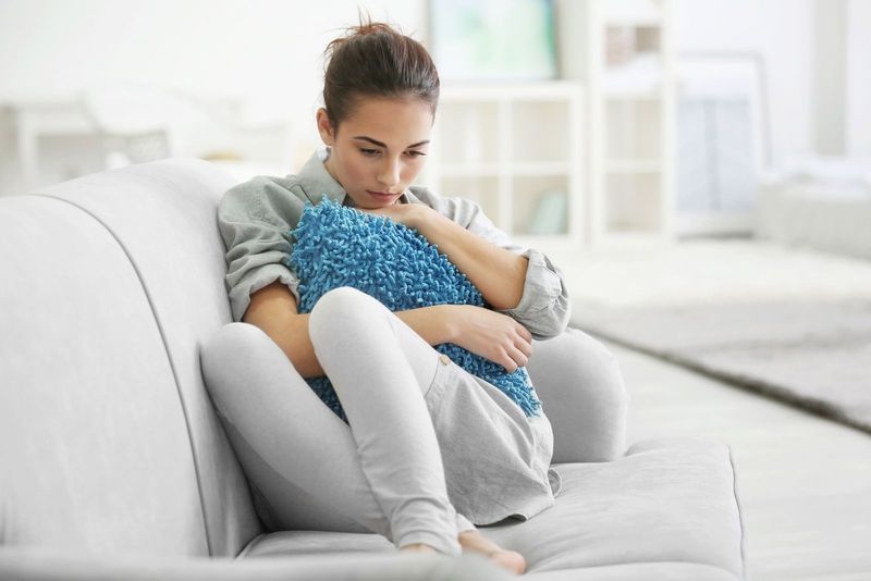 Depressive junge Frau sitzt mit den Füßen in der grauen Couch und umarmt das Kissen im Wohnzimmer
