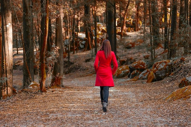 Junge Frau geht alleine auf einem Waldweg davon und trägt einen roten Mantel