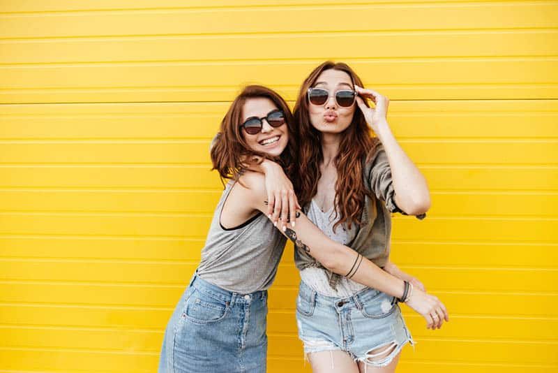 Fröhliche Freundinnen tragen Sonnenbrillen und posieren vor der Kamera mit gelbem Hintergrund