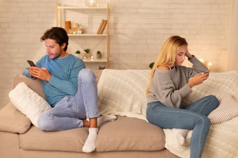 Ein Paar sitzt gelangweilt auf dem Smartphone und sitzt am gegenüberliegenden Ende der Couch im Wohnzimmer