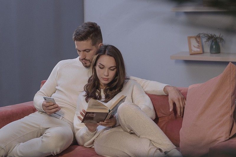 Eine Frau, die ein Buch liest, und ein Mann, der sein Smartphone benutzt, sitzen dicht beieinander auf der Couch
