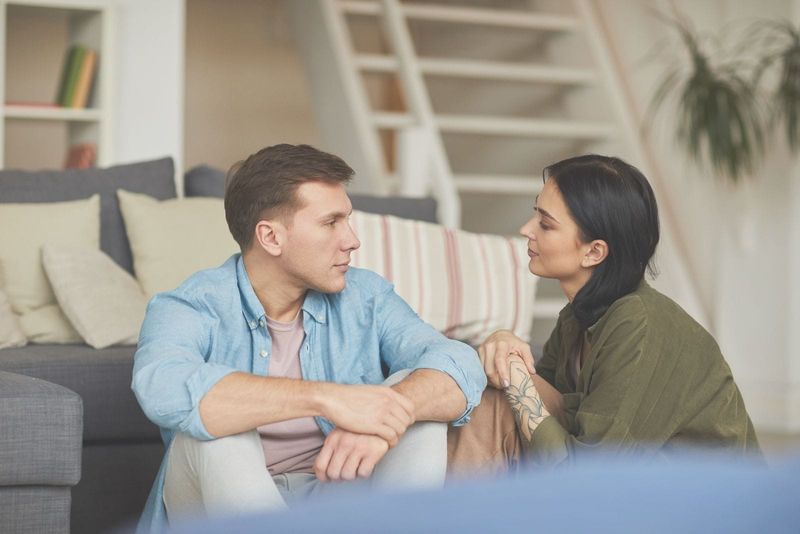 Modernes junges Paar, das aufrichtig miteinander redet, während es bequem auf dem Boden neben dem Sofa im Wohnzimmer sitzt